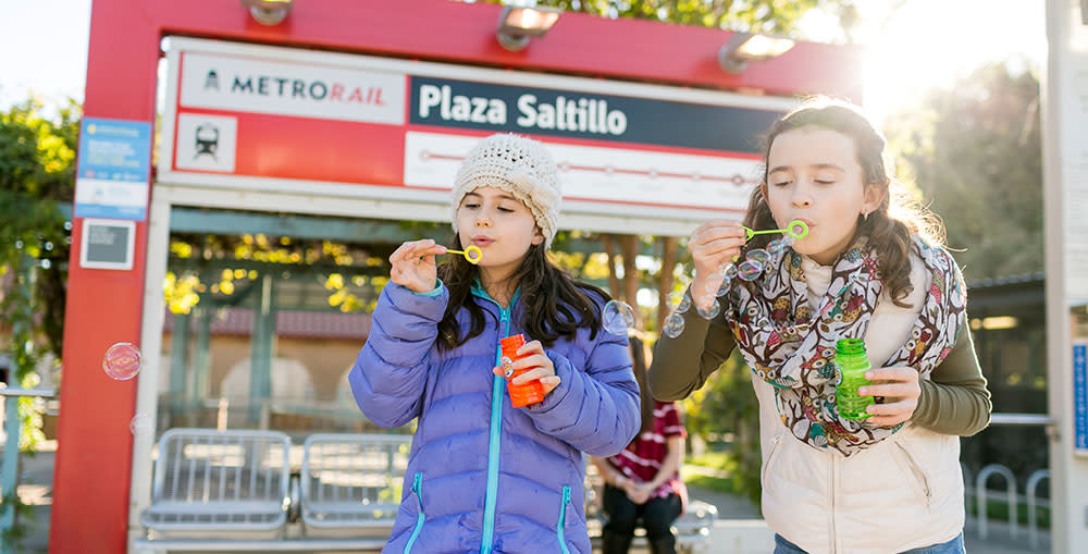 Two girls blowing bubbles in front of a MetroRail sign.