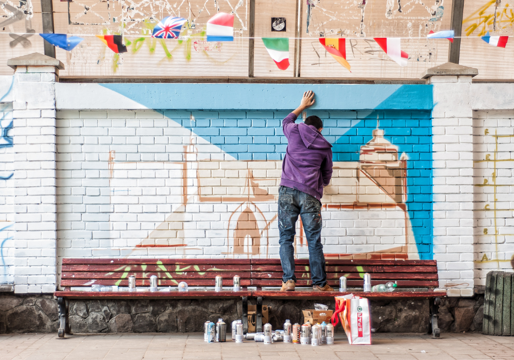 An artist standing on a bench starting to paint a mural on a brick wall.