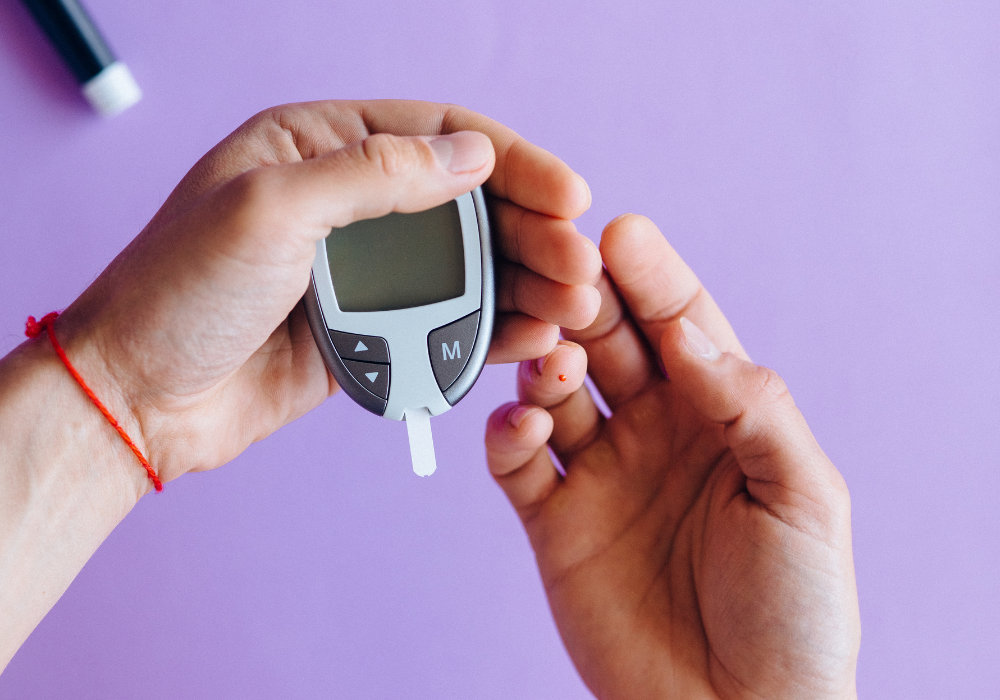 A person's hands using a glucometer to check their blood sugar with a lavender background