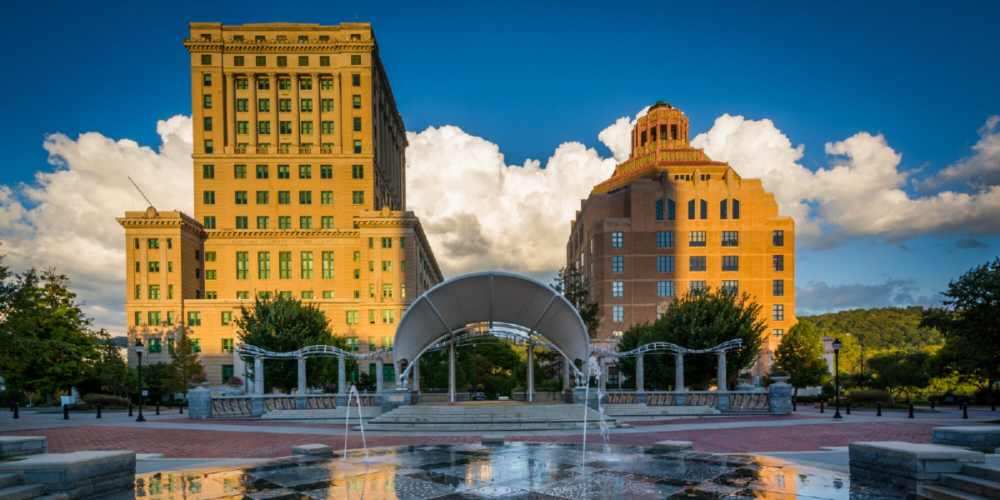 Scenic view of Asheville City Hall and the Buncombe County Courthouse, framed by a vibrant blue sky with fluffy white clouds. In the foreground, the iconic Splasheville fountain and an arched pavilion are surrounded by greenery and public space.