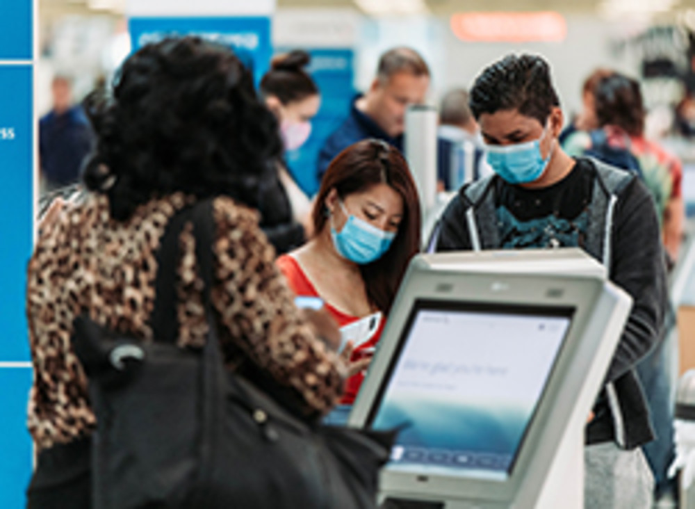 A woman using a digital kiosk to procure her flight ticket, there is a man and a woman both masked using another digital kiosk in the background