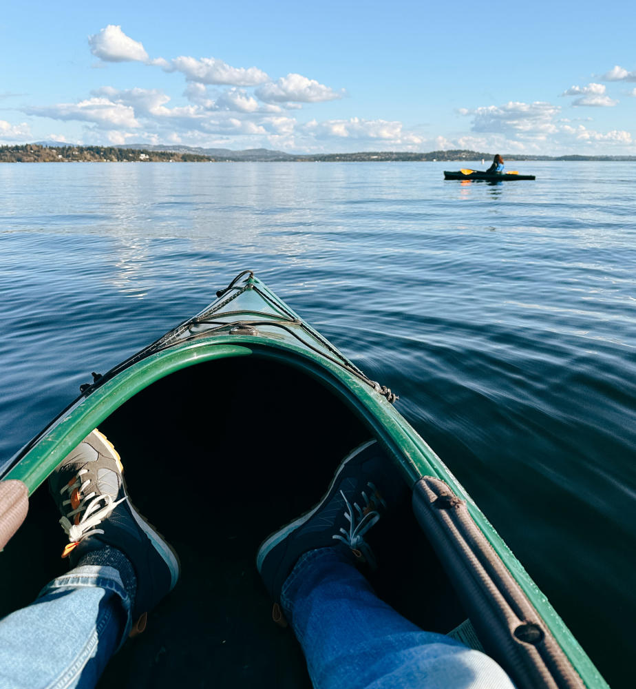 A pair of legs inside a kayak in the foreground with another kayaker floating on water in the background.