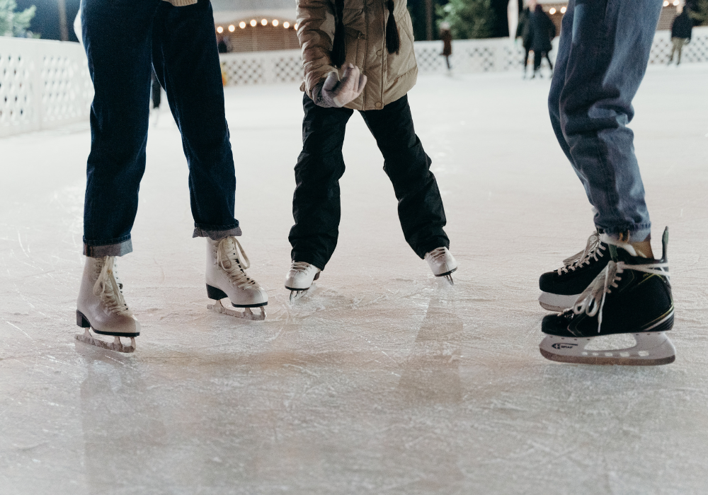 “Three people, bundled up in winter clothes, stand on ice wearing ice skates. The photo is taken from the waist down.
