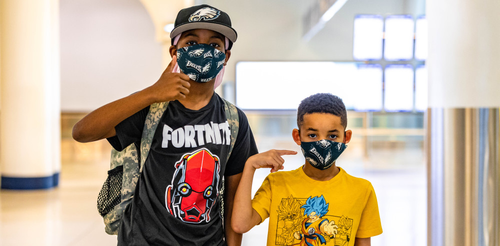 Two boys wearing their face masks give a thumbs up and point to their masks while traveling through the Charlotte airport. 