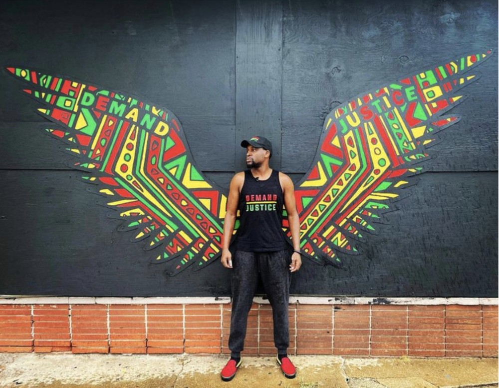 Artist Maxwell Emcays stands in front of a mural of wings.