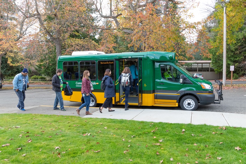 Une photo de six personnes faisant la queue pour monter à bord d'un bus près d'un parc public. Ils sont vêtus avec désinvolture de vestes, de jeans et de sacs à dos.