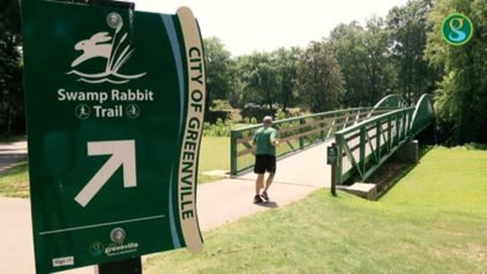 sign for Swamp Rabbit trail with a pedestrian crossing a bridge in the background