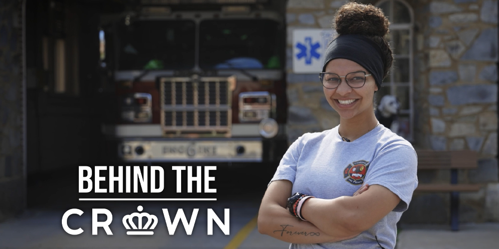 female firefighter smiling with arms crossed, fire truck, Behind the Crown logo
