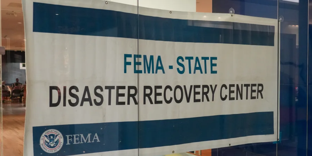 Sign for a FEMA-State Disaster Recovery Center, with FEMA branding, displayed on a window, indicating an active recovery site providing assistance to residents impacted by recent disasters.