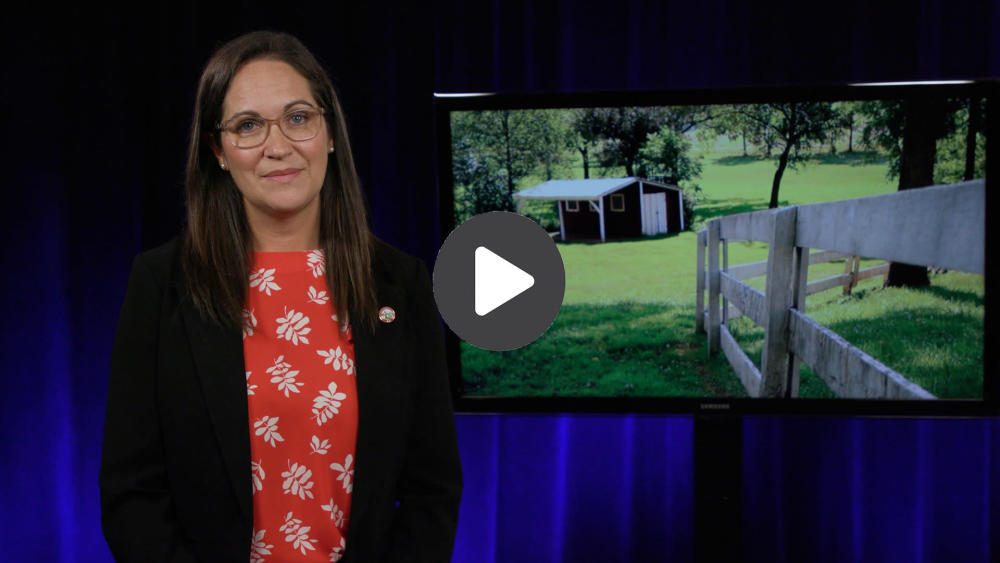 A woman standing inside next to a monitor. On the monitor is a photo of a farm house and fence.