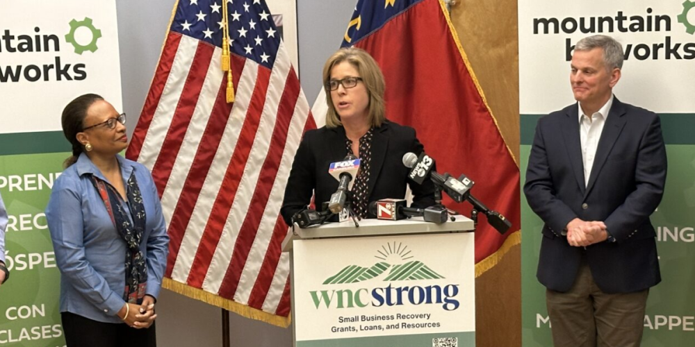 HUD Secretary Adrianne Todman, Asheville Mayor Esther Manheimer, and North Carolina Governor Josh Stein stand at a podium during a press conference at Mountain BizWorks, announcing federal disaster recovery funds for Asheville and North Carolina families affected by Hurricanes Helene and Milton.