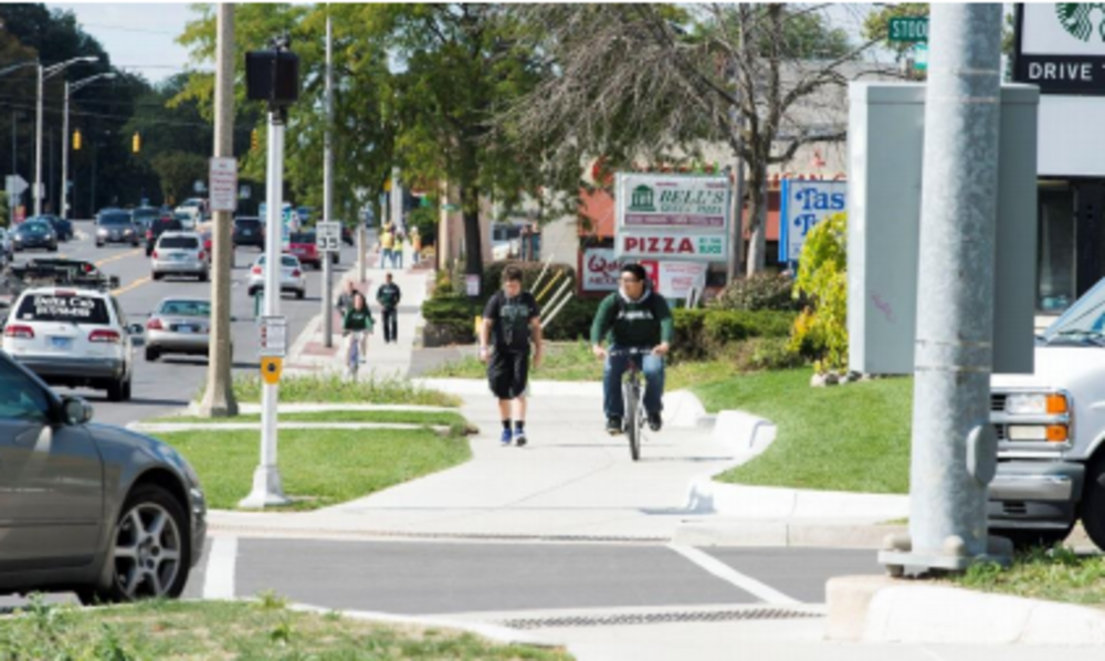 In the foreground, one person walking and one person riding a bike on a multi-use sidepath with a turning vehicle at the upcoming intersection