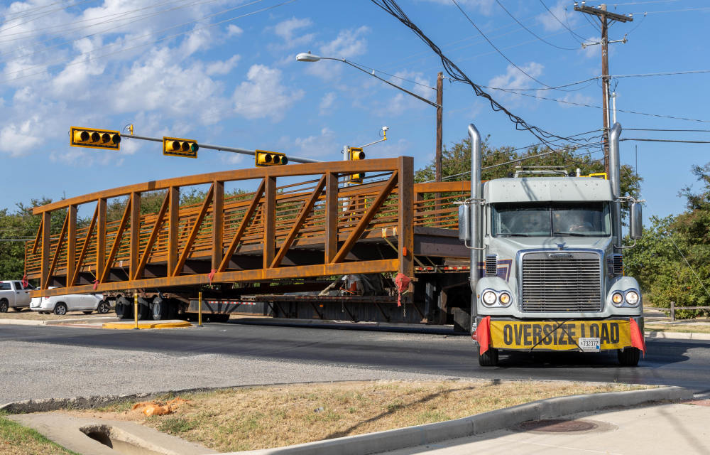 Image of a freight truck with a yellow sign that reads "oversize load," delivering the replacement pedestrian bridge to be installed at Roy G. Guerrero Park.