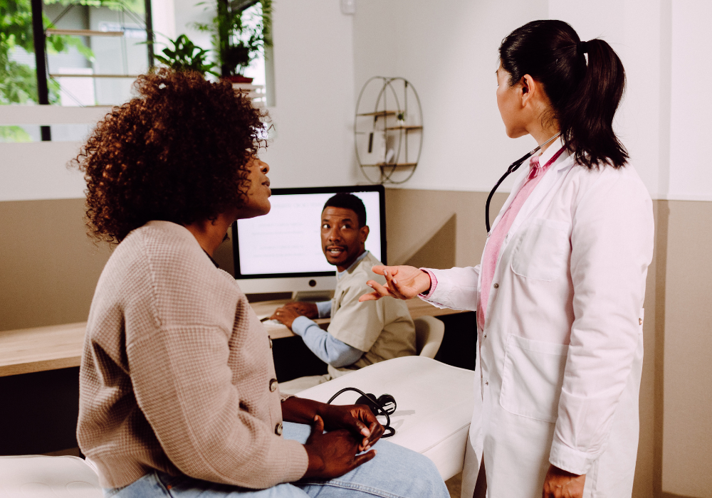 Doctor in white coat standing next to female patient sitting on a bed. The doctor is talking to a male medical staff member seated nearby at a computer.