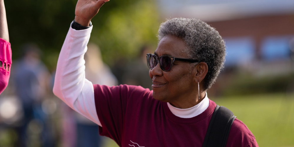 City Manager Debra Campbell smiles warmly and gives a high five during an outdoor community event, wearing a maroon shirt and sunglasses, with a sunny backdrop of blurred greenery.